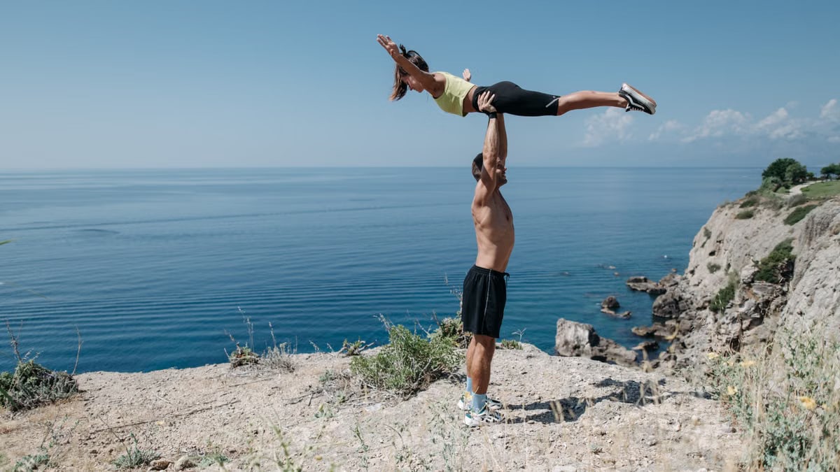 A couple performs acrobatic exercise on a scenic ocean cliff, embodying fitness and adventure.
